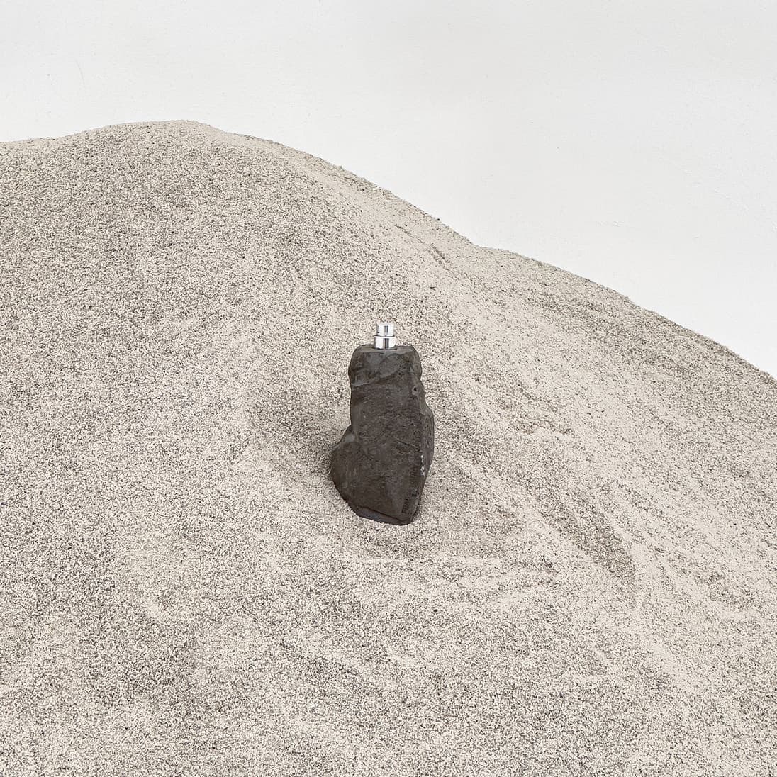 A dark, boulder-shaped concrete perfume bottle with a silver spray nozzle, half buried in a mound of sand against a white wall