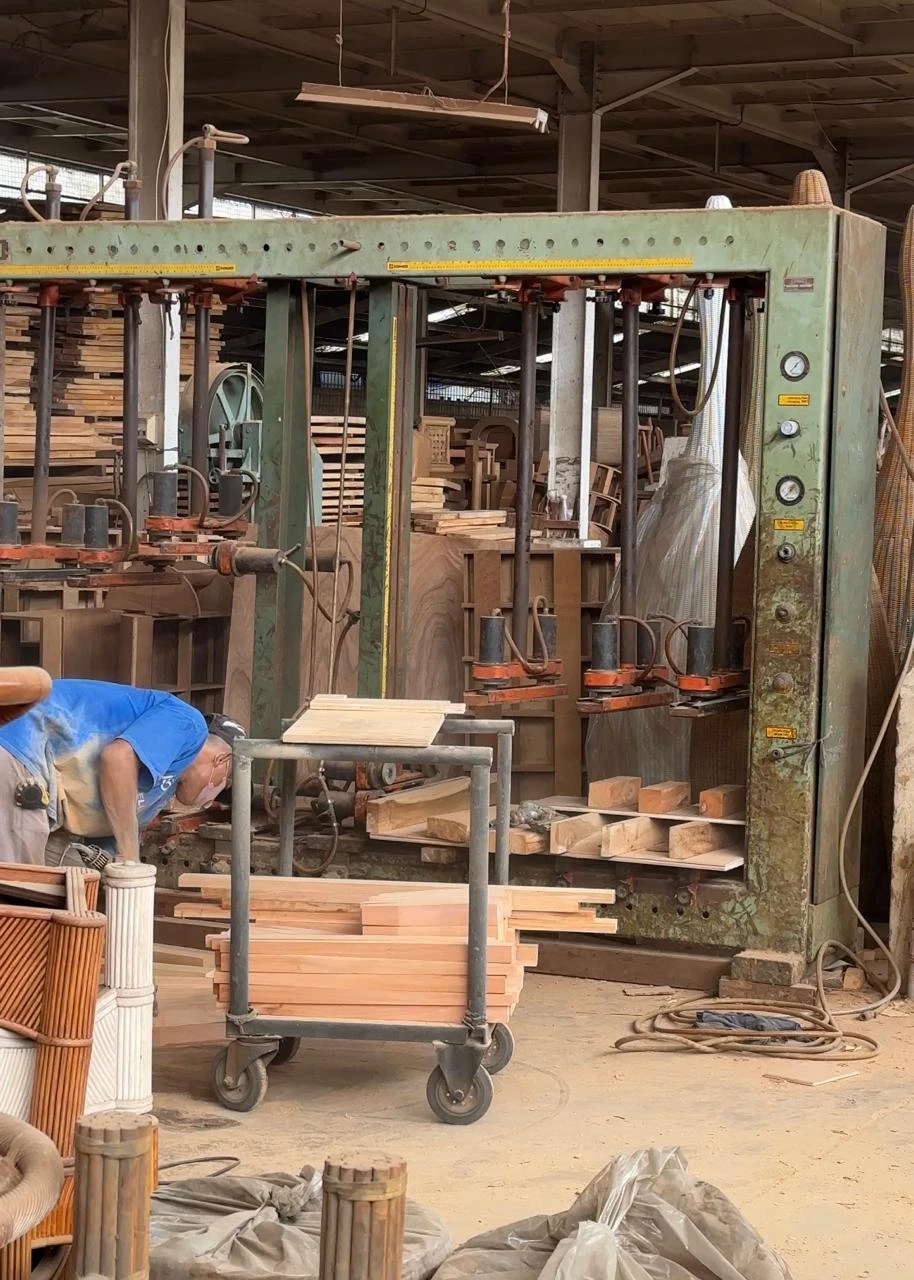 A wood factory floor under a steel roof: a large green clamping press with orange cylinders holding cut boards, a masked worker in a blue shirt bending over at the left, a cart stacked with timber in front, and rattan furniture parts in the foreground